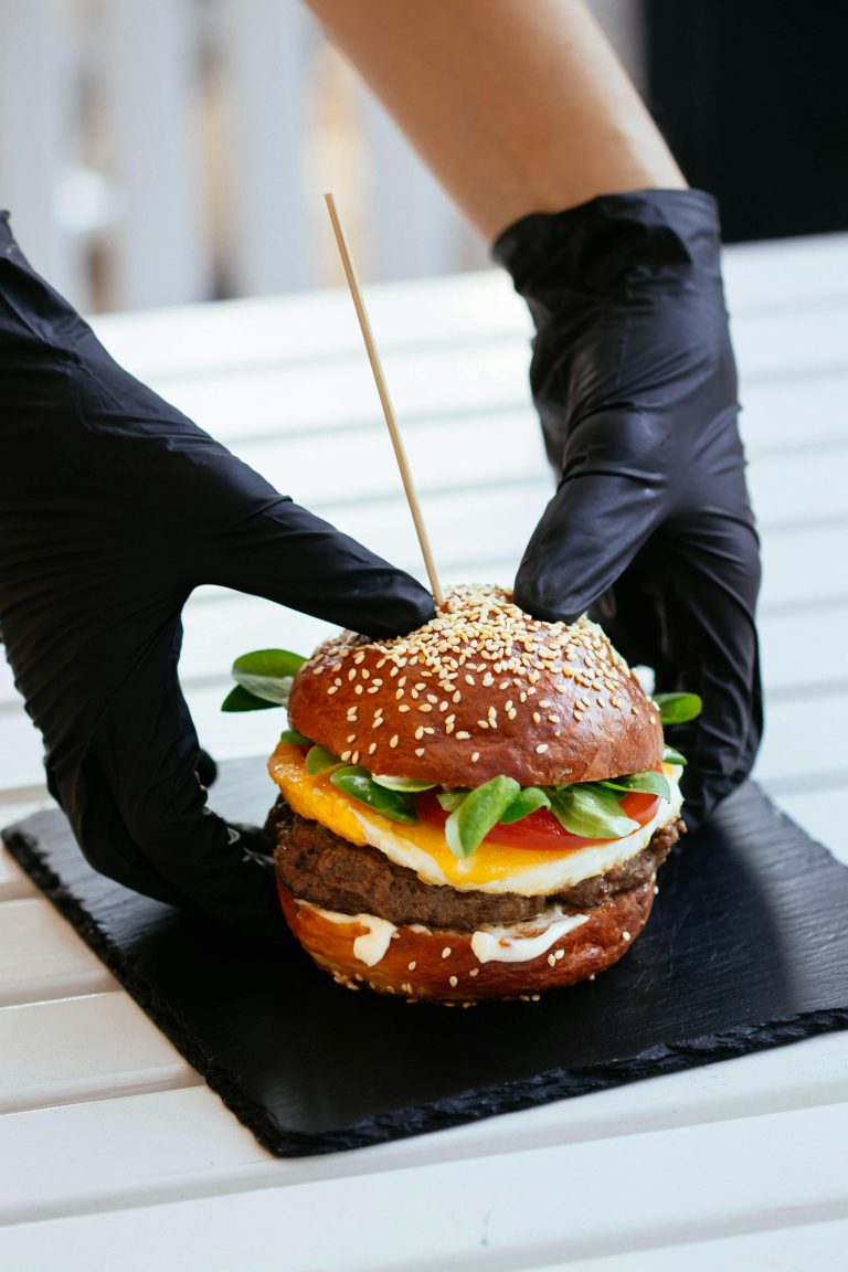 A delicious gourmet burger with sesame bun and fresh vegetables, held by gloved hands on a slate plate.