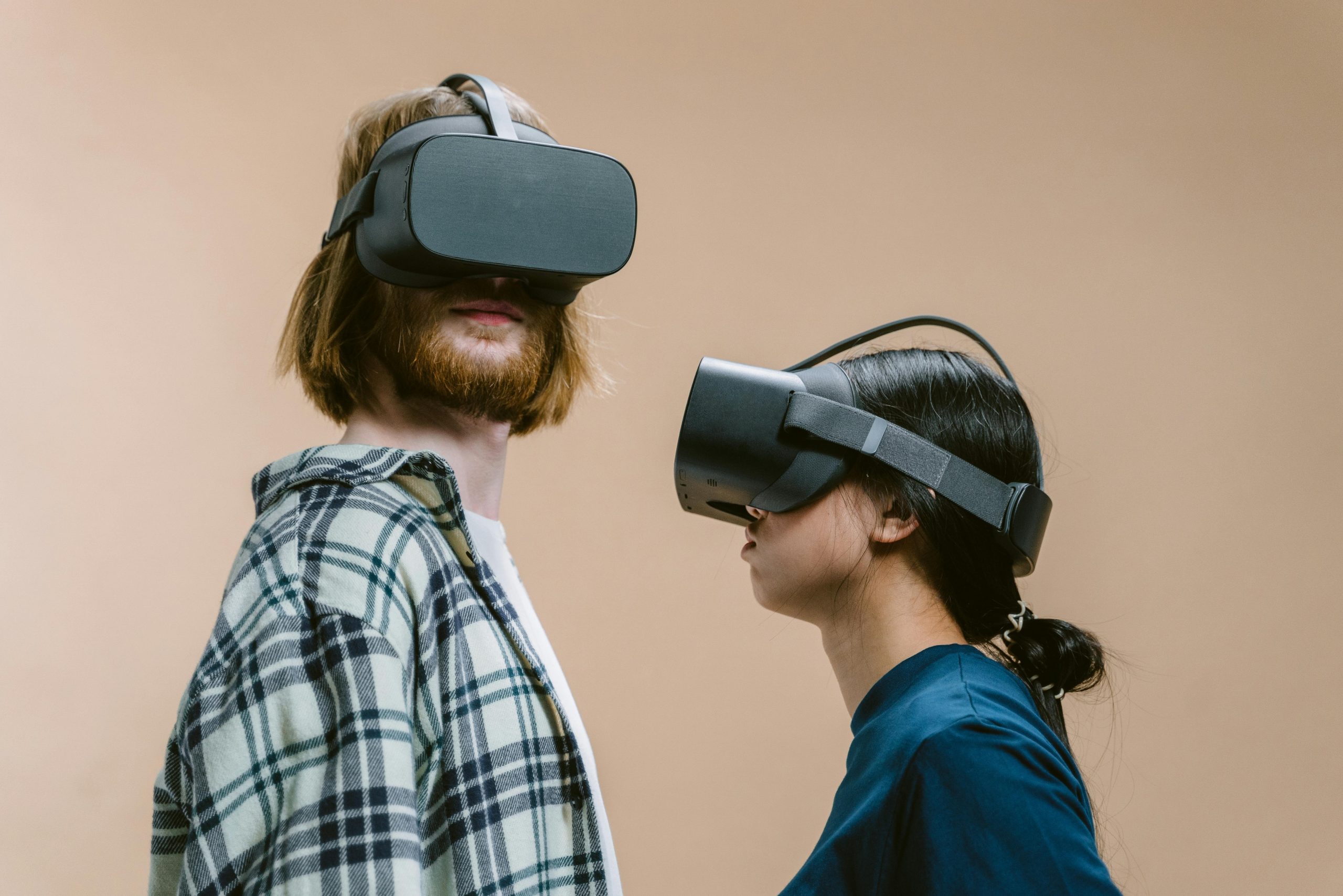 A man and woman wearing VR headsets, exploring virtual reality indoors with beige background.